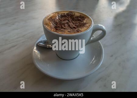 Aus nächster Nähe sehen Sie einen Cappuccino, Schaum und Schokoladenchips, serviert auf einer weißen Tasse über einer Untertasse, mit einem Teelöffel auf einem Marmortisch. Stockfoto