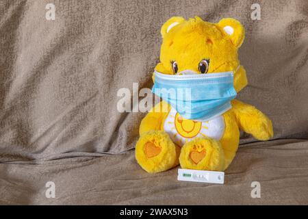 Nahaufnahme eines gelb-weiß gefärbten Teddybären auf einem Sofa mit blauer Gesichtsmaske während der COVID-19-Pandemie mit positivem Antigenschnelltest. Stockfoto