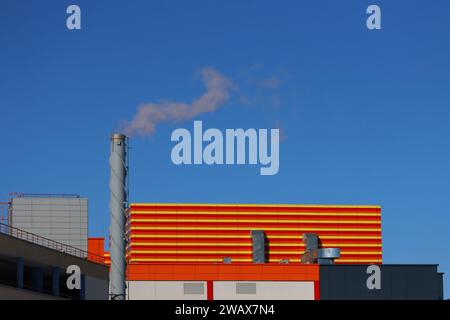 Schornstein mit Rauch, der in einer Fabrik rauskommt. Ein rauchender Kamin eines orangen Industriebaus vor blauem Himmel. Stockfoto