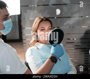 Eine Frau in einem Zahnarztstuhl schaut in den Spiegel auf ihre geheilten weißen Zähne und ein schneeweißes Lächeln. Hübsche Frau lächelt im Zahnarztstuhl, Doktor hält Stockfoto