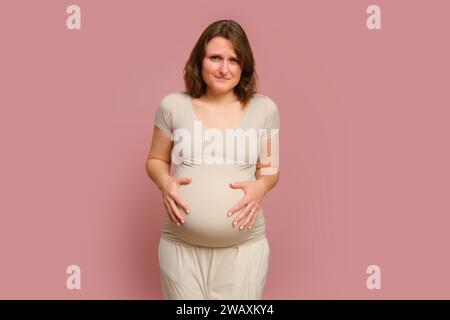 Eine schwangere Frau hält sich an einem schmerzenden Magen auf einem rosa Hintergrund des Studios fest. Schwangerschaft bei einer Frau mit Bauch, Kopierraum Stockfoto