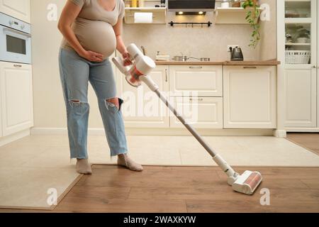 Die schwangere Frau benutzt einen kabellosen Staubsauger, um die Küche zu reinigen. Die werdende Mutter putzt gerne die Küche mit einem kabellosen Staubsauger. Stockfoto