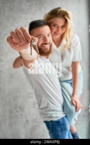 Ein fröhlicher Mann, der seine Frau mit einem Lächeln auf den Lippen hielt und dabei die Schlüssel zeigte. Glückliches Paar Immobilienkäufer demonstrieren Schlüssel von neuem Haus. Mit den Tasten auf die männliche Hand fokussieren. Stockfoto