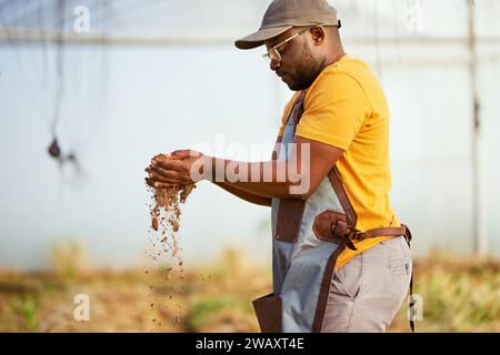 Schwarzer Bauer lässt Boden durch seine Hände fallen, im Hintergrund ein Gewächshaus. Stockfoto