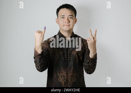 Asiatischer Mann mit Batiktuch zeigt seinen kleinen Finger mit violetter Tinte und wählt nach Pemilu oder Indonesien die Nummer zwei Stockfoto