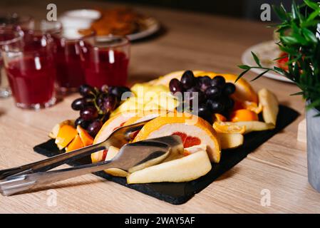 Eine lebhafte Auswahl an Ananasscheiben, Trauben und Orangen auf einem Schieferbrett mit Gläsern roter Getränke im Hintergrund. Stockfoto