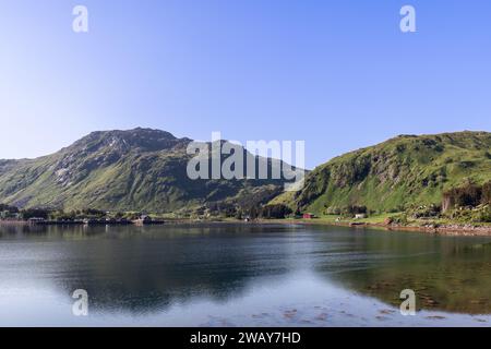 Ein ruhiger Morgenblick auf einen stillen Fjord in Nordnorwegen, mit üppigen grünen Hügeln und einer kleinen Ansammlung roter Häuser am Ufer. Lofoten Isla Stockfoto