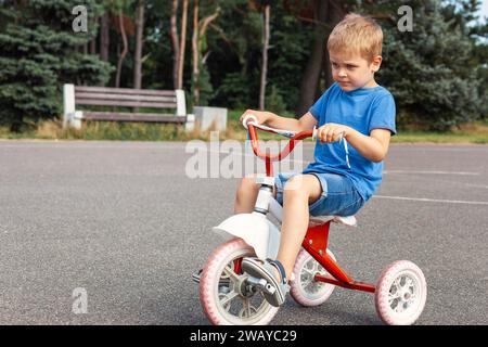 Ein kleiner Junge in blauen Kleidern, sehr konzentriert, fährt ein rotes Dreirad im Stadtpark. Stockfoto