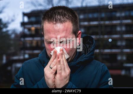 Bayern, Deutschland - 6. Januar 2024: Ein kranker Mann mit Erkältung bläst sich mit einem Papiertaschentuch die Nase *** ein kranker erkälteter Mann putzt sich mit einem Papiertaschentuch die Nase Stockfoto