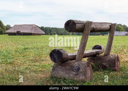 Eine Holzbank aus großen Baumstämmen steht in den breiten grünen Feldern des Dorfes, sie ist für Entspannung und Meditation bestimmt, mit alten Landhäusern Stockfoto