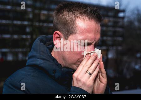 Bayern, Deutschland - 6. Januar 2024: Ein kranker Mann mit Erkältung bläst sich mit einem Papiertaschentuch die Nase *** ein kranker erkälteter Mann putzt sich mit einem Papiertaschentuch die Nase Stockfoto