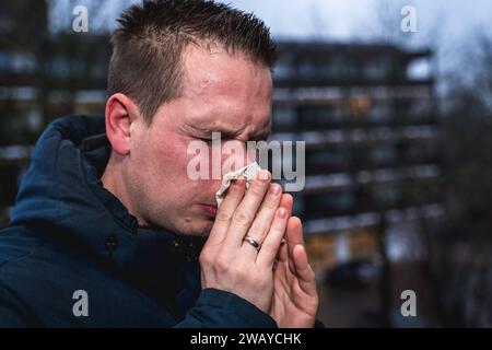 Bayern, Deutschland - 6. Januar 2024: Ein kranker Mann mit Erkältung bläst sich mit einem Papiertaschentuch die Nase *** ein kranker erkälteter Mann putzt sich mit einem Papiertaschentuch die Nase Stockfoto