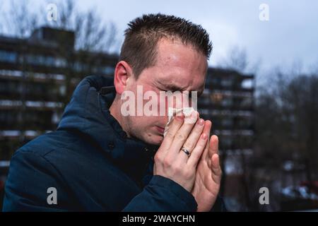 Bayern, Deutschland - 6. Januar 2024: Ein kranker Mann mit Erkältung bläst sich mit einem Papiertaschentuch die Nase *** ein kranker erkälteter Mann putzt sich mit einem Papiertaschentuch die Nase Stockfoto