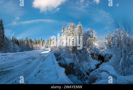 Sekundäre Landschaft alpine Straße zu abgelegenen Bergdörfern durch verschneiten Tannenwald, Schneeverwehungen und Holzzaun auf dem Weg Stockfoto