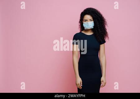 Feierliche Frau in schwarzem Kleid und Gesichtsmaske, rosa Hintergrund. Stockfoto