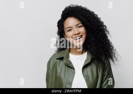 Lächelnde Frau in grüner Jacke über weißem Oberteil, weißem Hintergrund Stockfoto