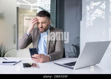 Besorgter junger Geschäftsmann, der im Büro am Tisch sitzt und enttäuscht auf den Telefonbildschirm blickt und seinen Kopf hält. Stockfoto