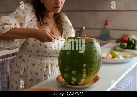 Details zu Wassermelone und beschnittener Ansicht einer Hausfrau im Sommerkleid, die reife, saftige Beeren aus biologischem Anbau schneidet und an der Küchenzeile steht. Gesundheit Stockfoto