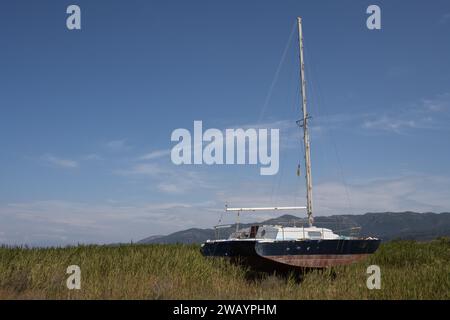 Alte verrostete verlassene Jacht auf der Akte mit hohem Gras. Berge im Hintergrund. Blauer Himmel mit hellweißen Wolken im Frühling. Roda, Korfu, G Stockfoto