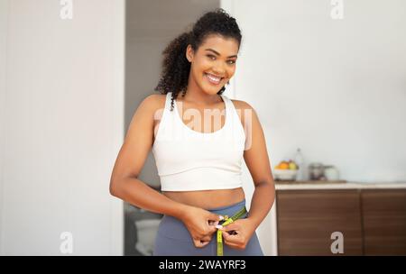 Sportliche junge Frau, die Taille misst, lächelt zu Hause in die Kamera Stockfoto
