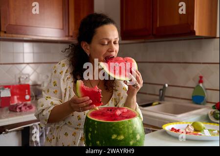 Lockiges Haar Multi ethnische hübsche Frau isst Wassermelone in rustikalen Wohnküche Innenraum Stockfoto