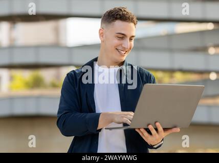 Ein glücklicher junger Mann, der draußen einen Laptop benutzt, in einer lässigen Kleidung Stockfoto