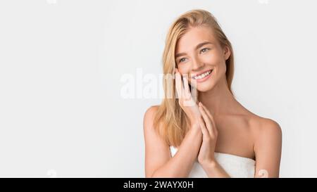 Schöne blonde Frau, in Handtuch gewickelt, berührt das Gesicht, weißes Studio Stockfoto