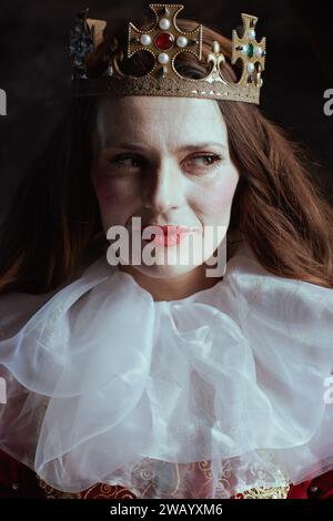 Nachdenkliche mittelalterliche Königin in rotem Kleid mit weißem Kragen und Krone auf dunkelgrauem Hintergrund. Stockfoto