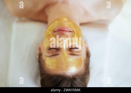 Gesundheitszeit. Obere Ansicht einer entspannten modernen Frau in Massagekabine mit goldener Kosmetikmaske auf dem Gesicht, das auf Massagetisch liegt. Stockfoto