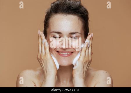Glückliche Frau mittleren Alters mit schäumendem Gesichtsreiniger, der Gesicht vor beigem Hintergrund wäscht. Stockfoto