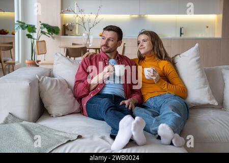 Zufriedenes, liebevolles Paar, das nach dem Job Tee getrunken hat und das Fenster auf einem gemütlichen Sofa in einem neuen Zuhause angeschaut hat. Stockfoto