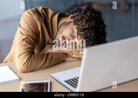 Geschäftsmann, der am Arbeitsplatz schläft, junger übermüdeter und überarbeiteter Mann, der auf dem Schreibtisch im Büro mit Laptop liegt, lange Arbeit bis spät, Abgabetermin am Abend. Stockfoto