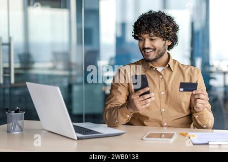 Geschäftsmann im Büro mit Telefon und Bankkreditkarte in Händen, Mann verwendet App auf Smartphone, um im Online-Shop zu kaufen, zufriedener Mitarbeiter im Büro mit Laptop. Stockfoto