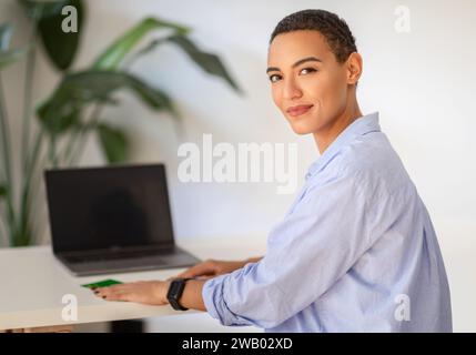 Eine professionelle junge Frau mit kurzen Haaren posiert selbstbewusst an einem Schreibtisch mit Laptop Stockfoto