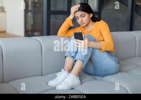 Besorgte junge indianerin mit Smartphone in den Händen, die auf dem Sofa sitzt Stockfoto
