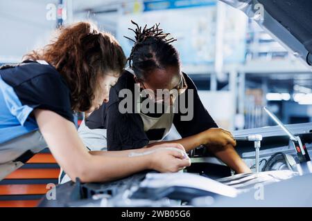 Ein Team von Frauen in der Autowerkstatt arbeitet zusammen an der Reparatur des Autos und diskutiert die besten Optionen. Teamwork-Mechaniker arbeiten an der Wartung defekter Fahrzeuge zusammen und prüfen auf beschädigten Kraftstofftank Stockfoto