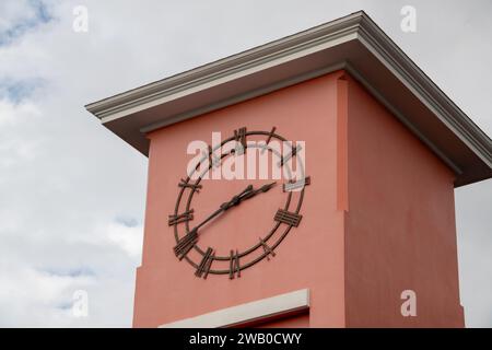 Eine analoge Außenuhr in einem rosafarbenen Turmgebäude mit einem cremefarbenen Flachdach. Die Hände lesen um 240 Uhr. Die Zahlen sind aus mir gemacht Stockfoto