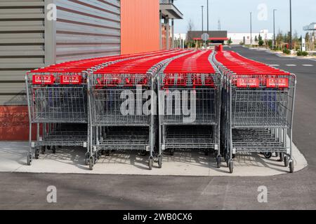 Reihen neuer Costco-Einkaufswagen aus Metall mit roten Griffen. Die Schubwagen befinden sich vor einer orangefarbenen und grauen Metallaußenwand eines Ladens Stockfoto