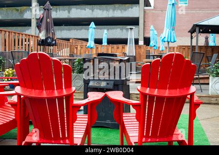 Der Lounge-Bereich im Freien eines Restaurants mit roten Adirondack-Stühlen, Heizungen, Sonnenschirmen, Tischen und Stühlen. Es gibt ein braunes Baldachin-Zelt und Holzzaun Stockfoto