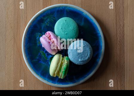 Vier französische Macarons; Rosa, Grün, Blau und Lila. Das elegante süße Dessert im Sandwich-Stil ist auf einem blauen Teller und Holztisch gestapelt. Stockfoto