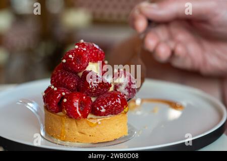 Mit Früchten gefüllte französische Pudding-Tarte. Das Topping enthält frische ganze rote Himbeeren auf einer Mandelkruste. Eine Hand hält eine kleine Gabel im Dessert Stockfoto