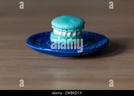 Ein einzelnes blaues Macaron gefüllt mit weißer Vanillecreme. Der französische Mandel-Macaron liegt auf einem kleinen blauen Teller und Holztisch. Stockfoto