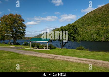 Picknicktische mit einer geschützten Metallabdeckung. Der Campingplatz liegt in der Nähe eines Flusses mit baumbewachsenen Hügeln, üppig grünem Gras und Flussufer. Stockfoto