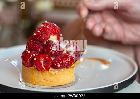 Mit Früchten gefüllte französische Pudding-Tarte. Das Topping enthält frische ganze rote Himbeeren auf einer Mandelkruste. Eine Hand hält eine kleine Gabel im Dessert Stockfoto