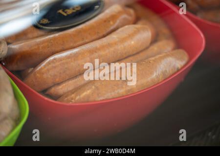 Eine rote Plastikschale gefüllt mit Taco-Würstchen oder Weinern in einem Vitrinenschrank mit schwarzem Schild und goldenen Buchstaben. Das ungekochte mexikanische Protein ist roh. Stockfoto