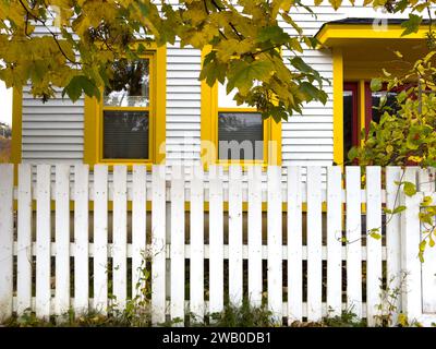 Das Äußere einer weißen cape Cod-Schrankwand, horizontale Seitenwand im Holzstil mit zwei hellgelben Zierfenstern. In der Nähe des Zauns ist ein Baum. Stockfoto