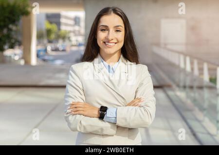 Selbstbewusste europäische Millennials-Geschäftsfrau mit gekreuzten Armen, die in urbaner Umgebung lächelt Stockfoto