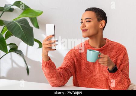 Die lächelnde Frau in einem orangefarbenen Pullover genießt einen Moment der Freizeit und hält eine Tasse Stockfoto