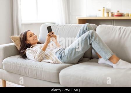Fröhliche junge Frau mit Smartphone in den Händen, die auf einem weichen Sofa liegt Stockfoto