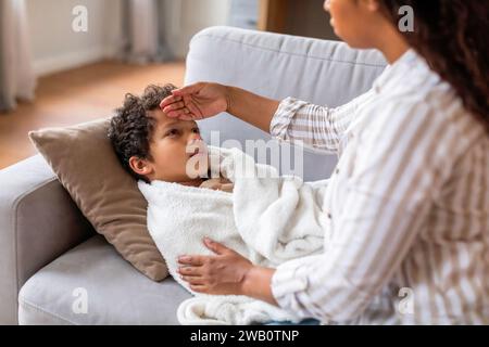 Die fürsorgliche schwarze Mutter überprüft die Stirn ihres Sohnes, während er auf der Couch liegt Stockfoto